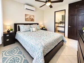 A bedroom with a bed, nightstands, a ceiling fan, an air conditioning unit, a bathroom visible through an open door, and a patterned rug.