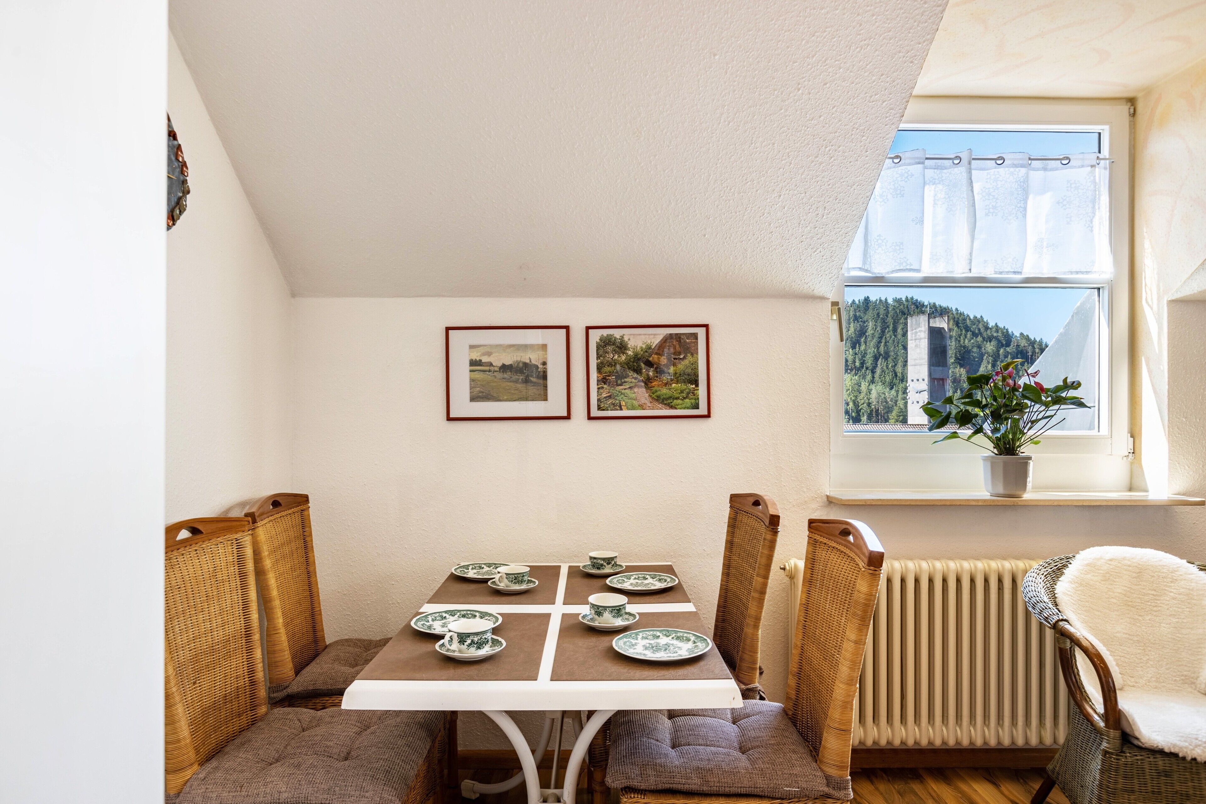 Un coin repas avec une table dressée pour quatre personnes, une chaise en osier, un radiateur et une fenêtre donnant sur une zone boisée.