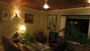 A cozy living room with a leather sofa, a coffee table with magazines, a lamp, and a television. There are framed pictures on the wall and a large window with curtains. - Hotel San Remo (Pinamar)