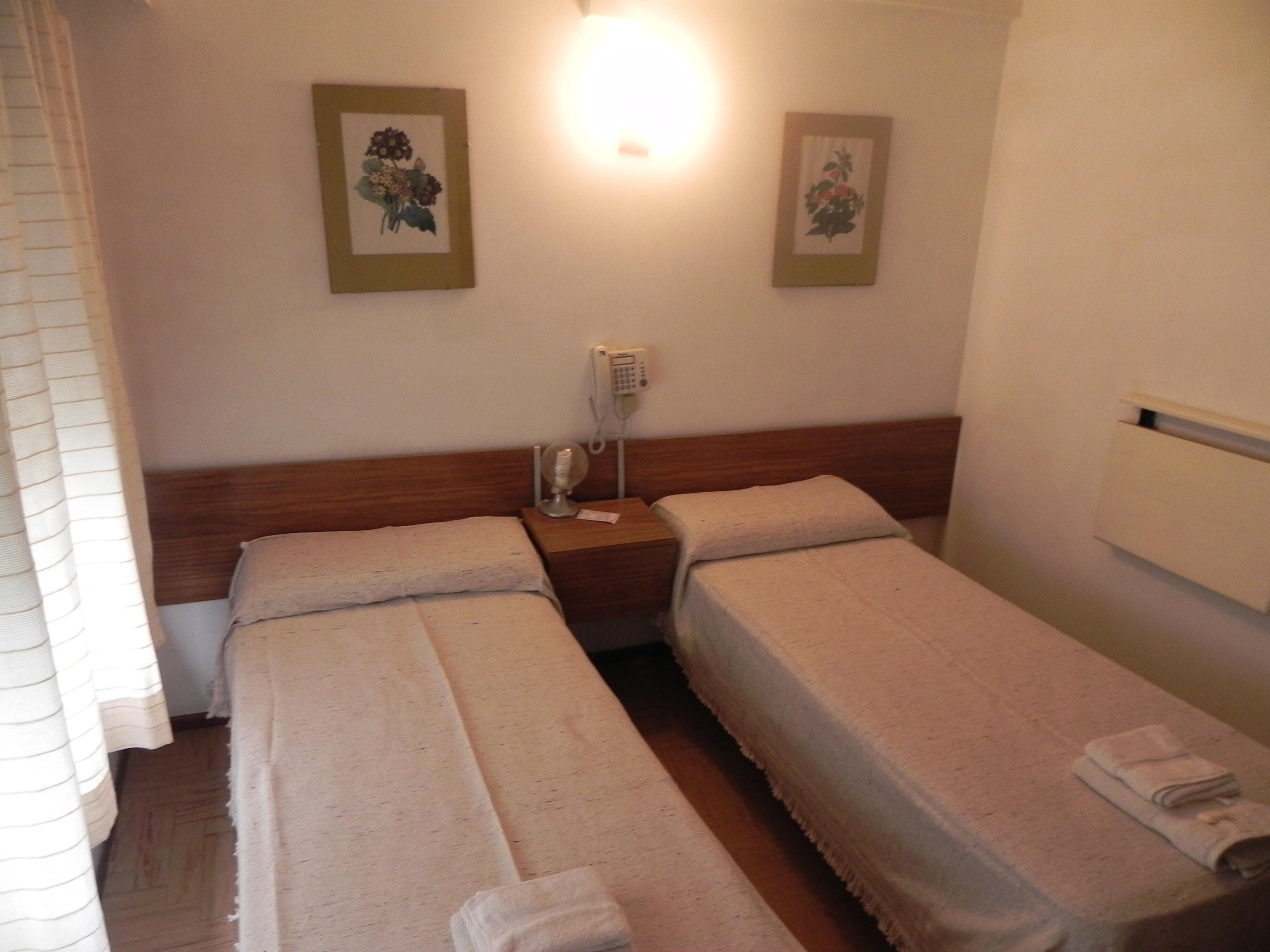 A hotel room with two single beds, a bedside table with a lamp, a phone, and two framed pictures on the wall.