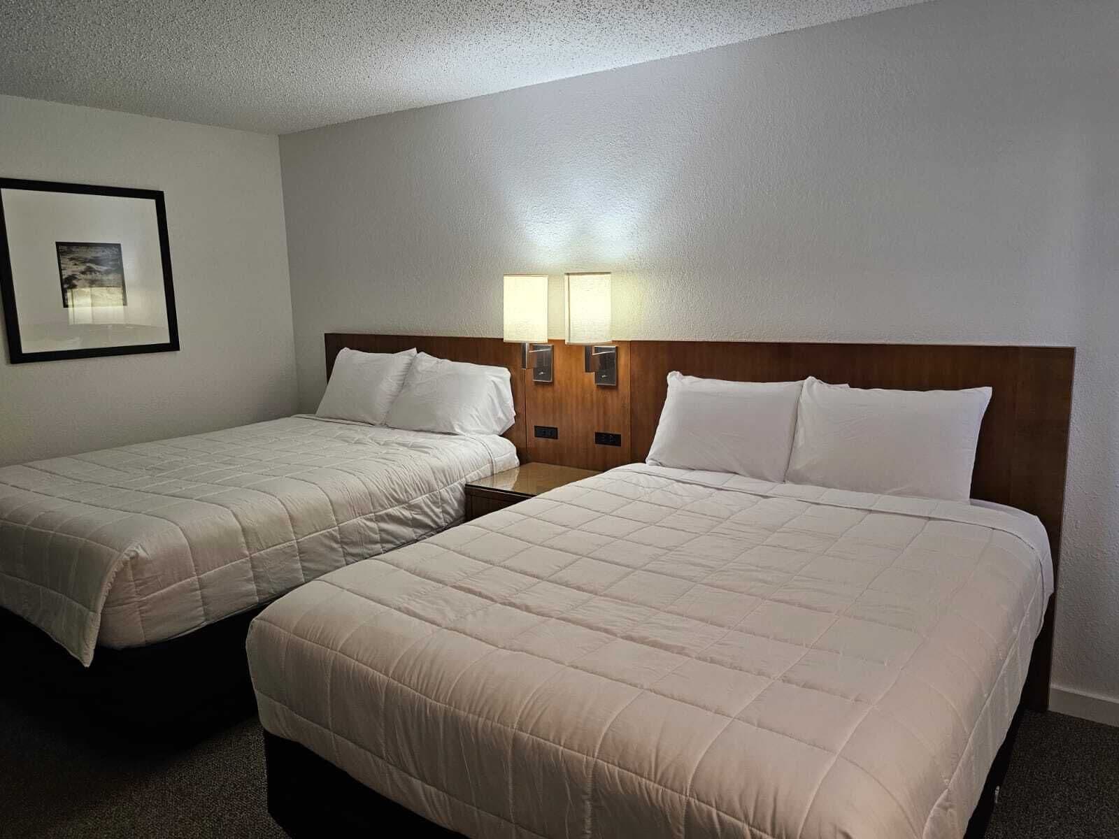 A hotel room with two beds, each with a wooden headboard and white bedding. There are two wall-mounted lamps on either side of the beds.
