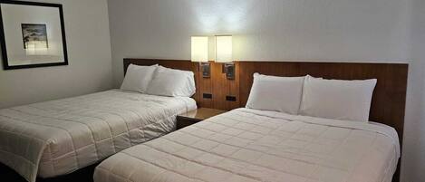A hotel room with two beds, each with a wooden headboard and white bedding. There are two wall-mounted lamps on either side of the beds.