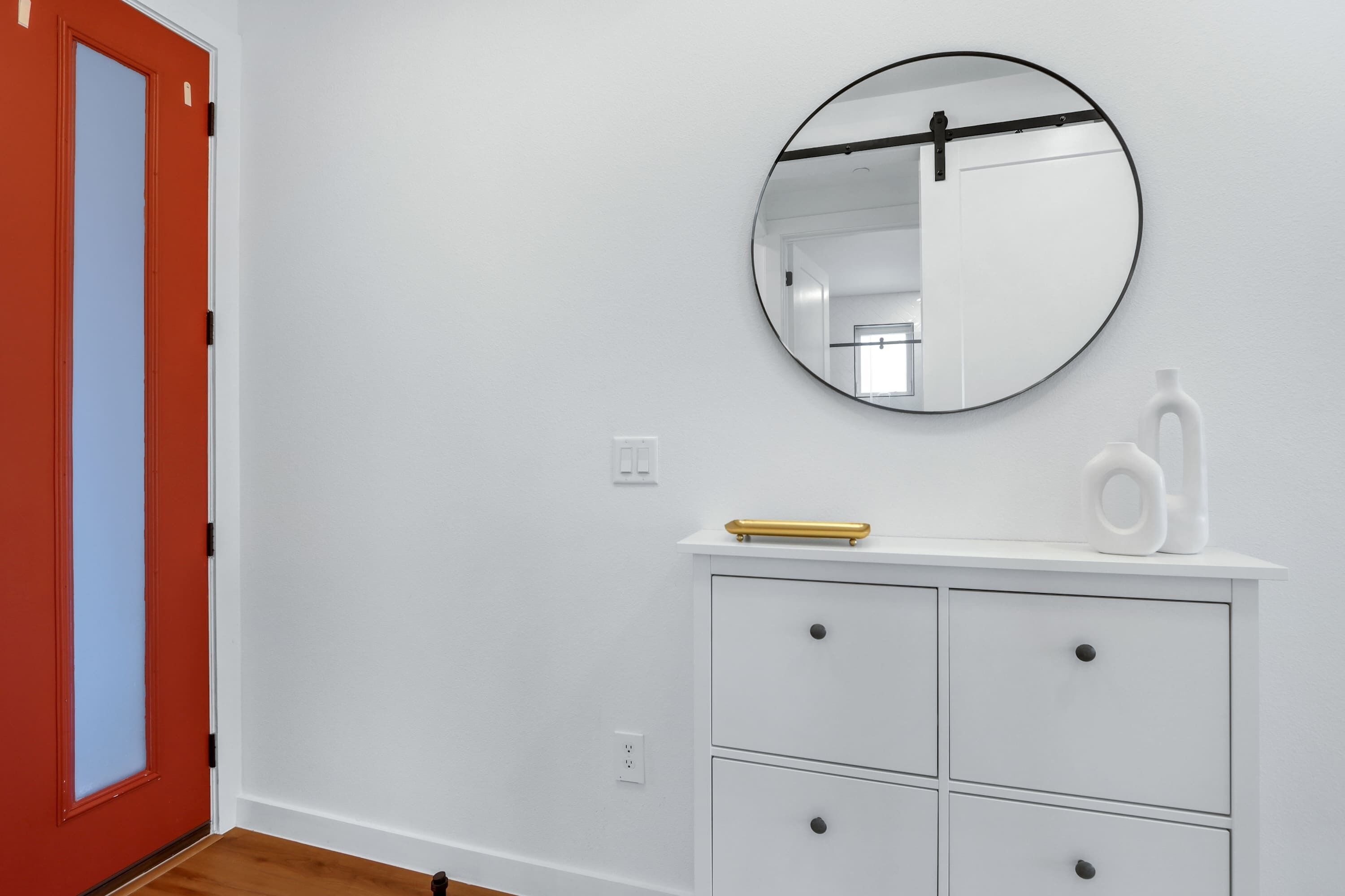 A room with a white wall, a round mirror, a white dresser with drawers, and a red door.