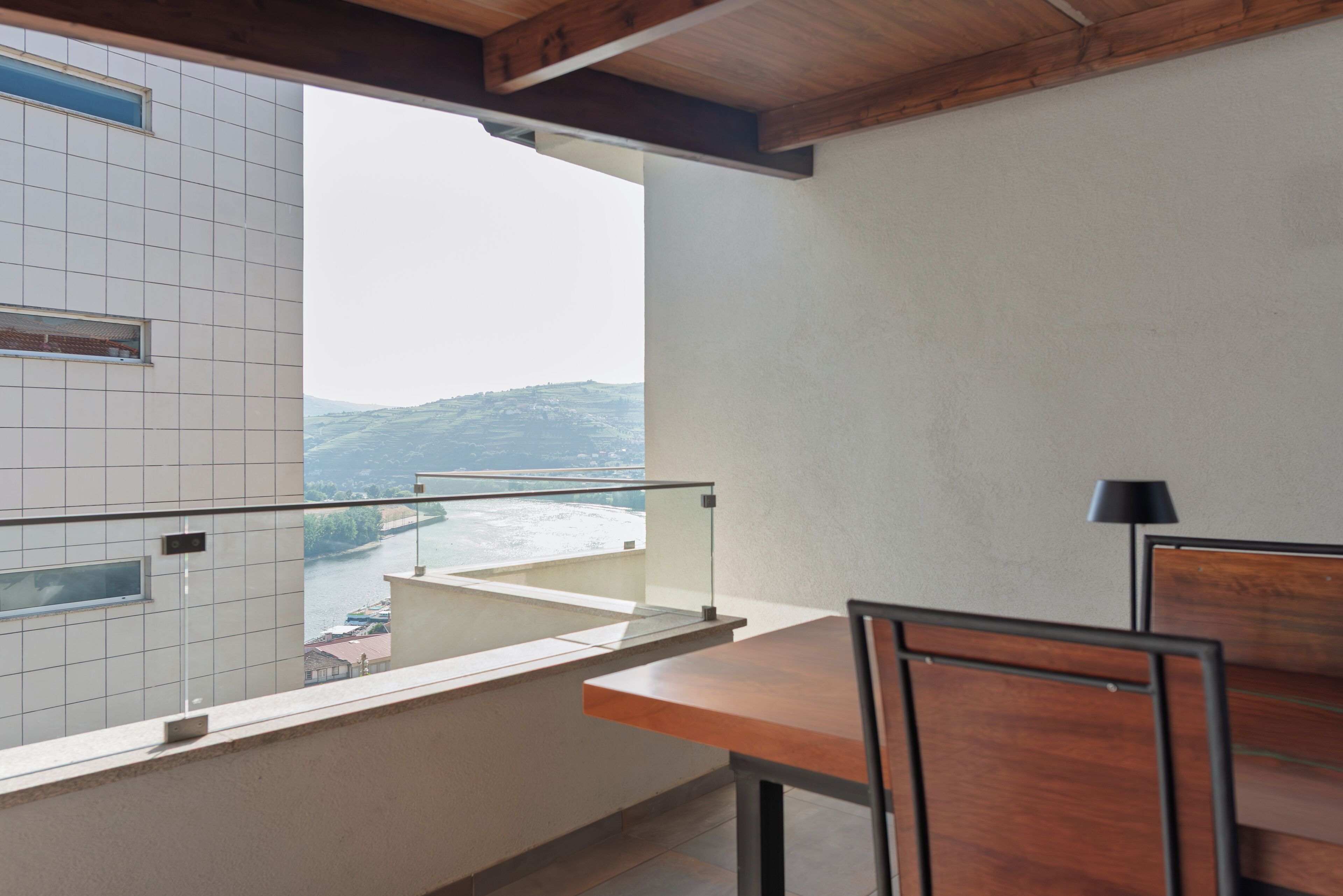 A balcony with a view of a river and hills, a wooden table, and a chair.