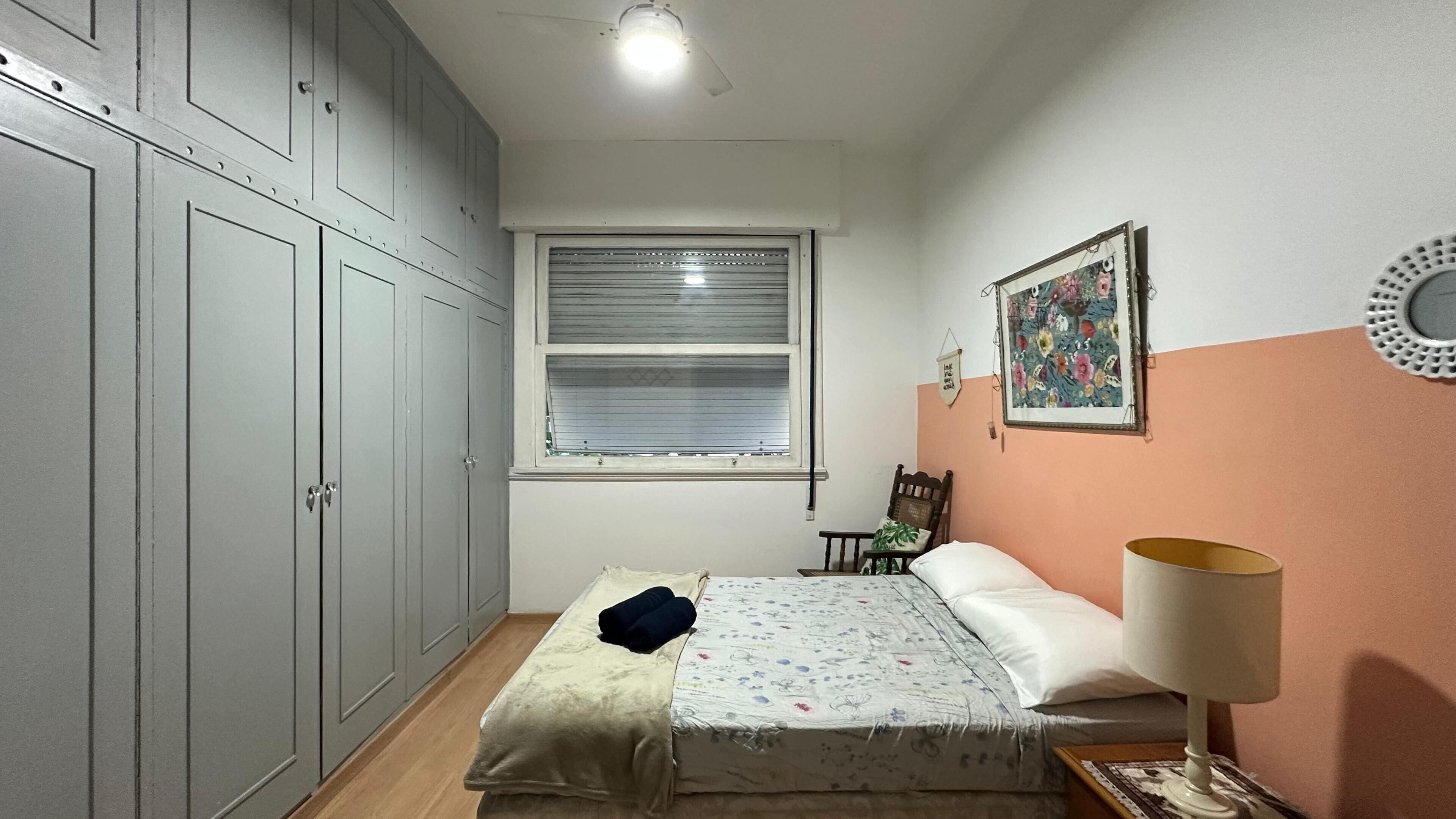 A bedroom with a bed, a nightstand, a window with blinds, a wardrobe, and a wall with a framed picture.