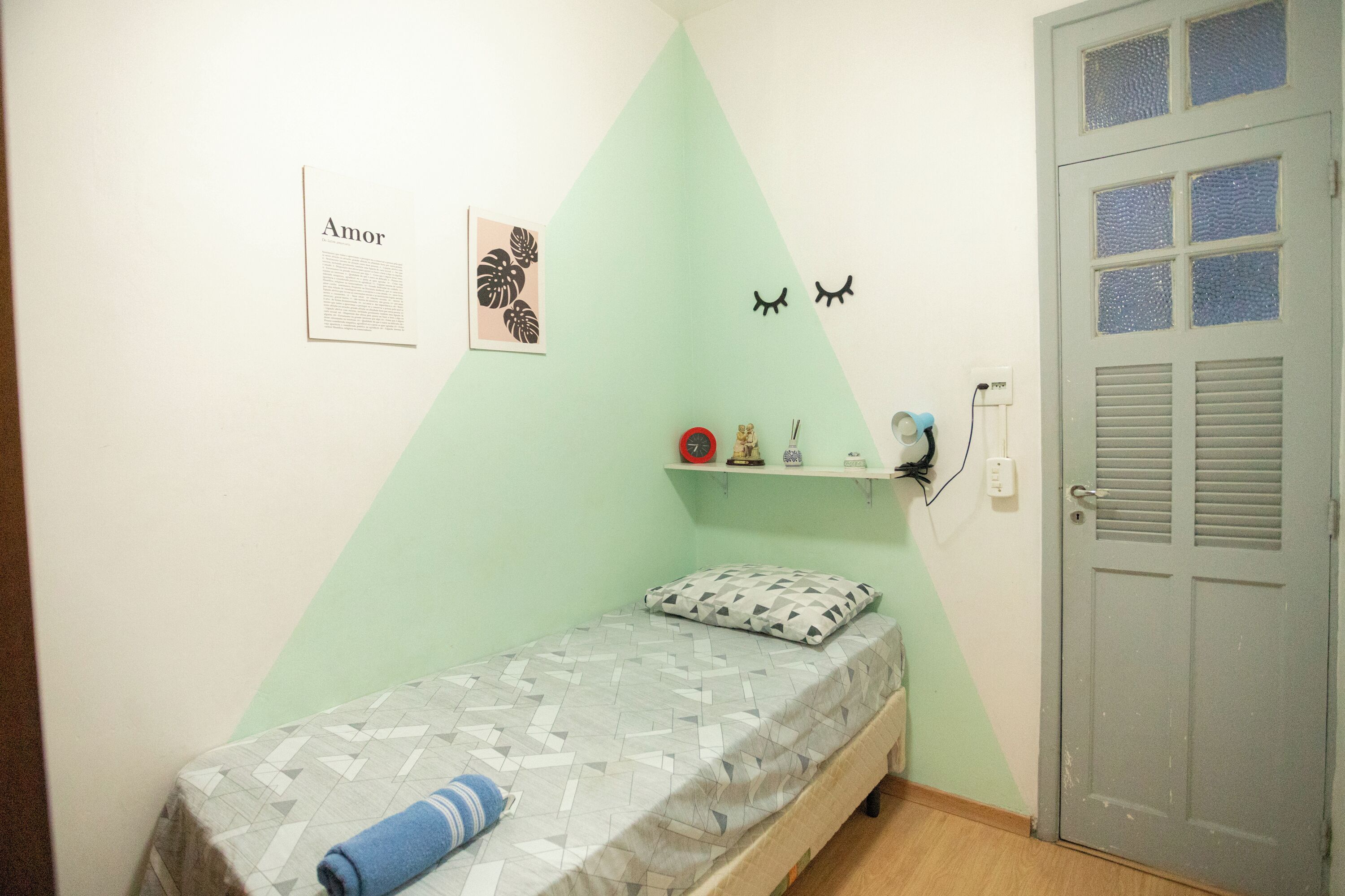 A small, neatly arranged bedroom with a single bed, a wall-mounted shelf with personal items, and a door with a window.