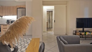 A modern living room with a sofa, a coffee table, and a television. There is a kitchen area with cabinets and a coffee machine. A hallway leads to a bathroom.