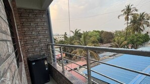 A balcony with a stone wall, a black trash bin, and a view of a blue-roofed building and palm trees. - 1 bhk comfy stay 1 (Pune)