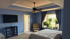 Un dormitorio con cama, una silla, televisor, ventilador de techo y ventana con cortinas.