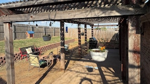 Un patio cubierto con una pérgola de madera, mobiliario exterior, luces colgantes y una maceta colgante con flores.