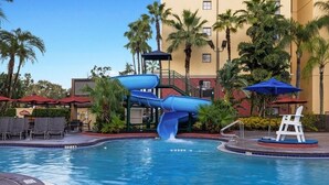 Una piscina con un tobogán acuático azul, rodeada de palmeras y sillas de descanso.