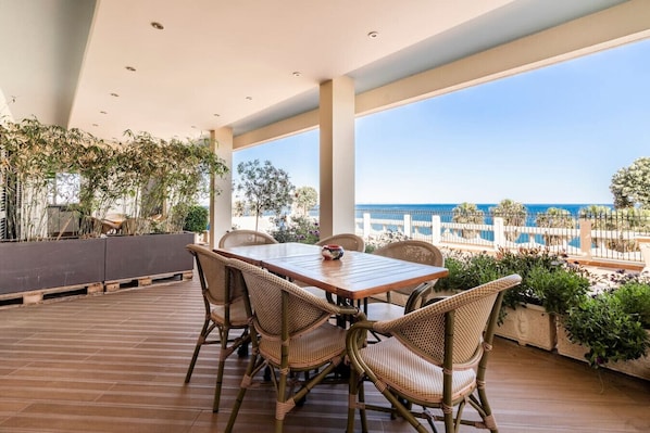 A covered outdoor dining area with a wooden table and wicker chairs, overlooking a pool and beach.