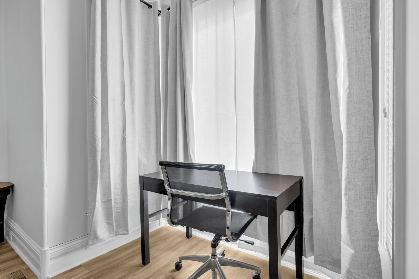 A modern office space with a black desk and office chair, white walls, and sheer curtains.