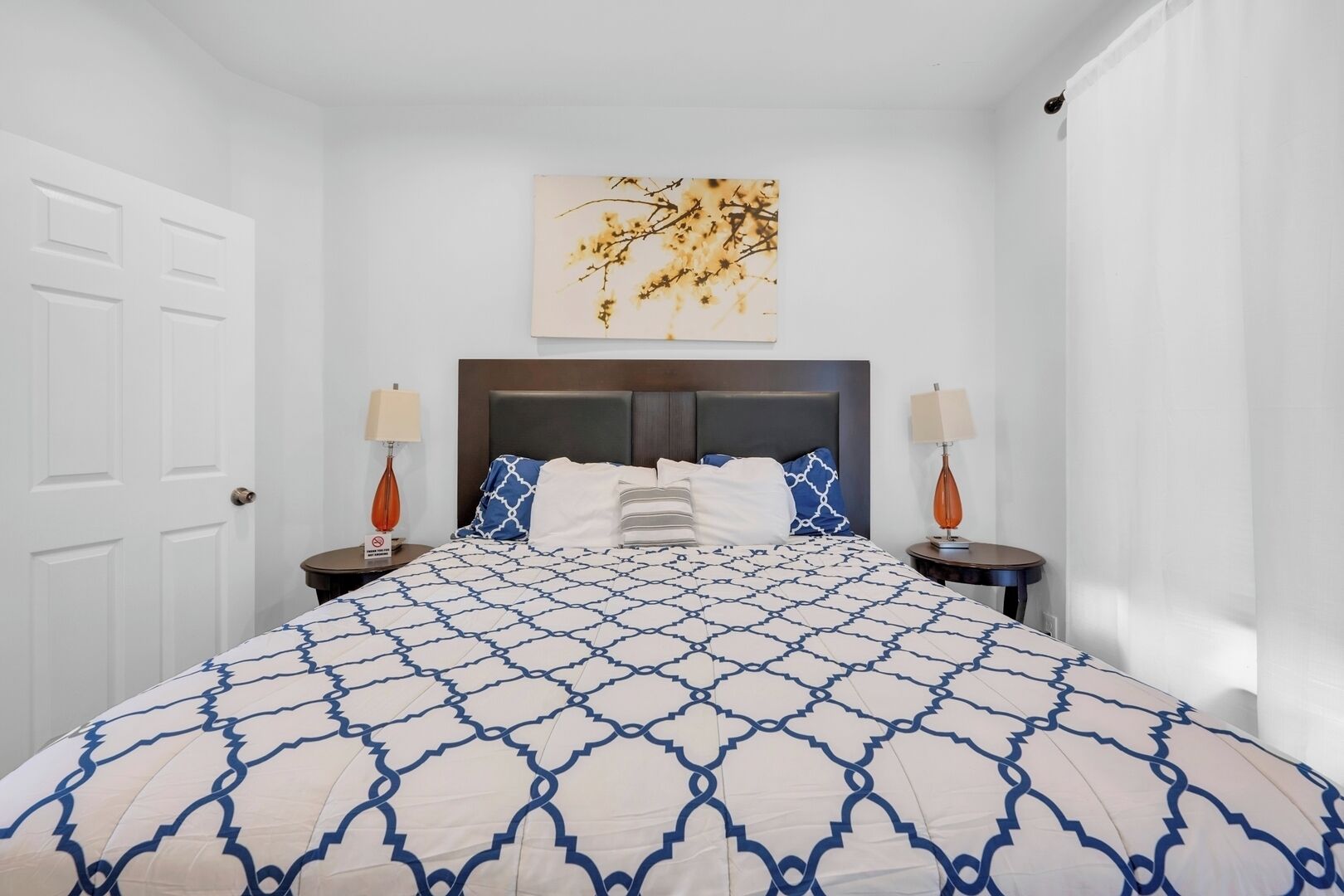 A neatly arranged bedroom with a large bed, two bedside tables, a wall-mounted artwork, and a white door.