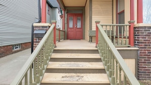 A front view of a two-story house with a red door, a front porch, and a staircase with green railings. - Spacious 3BR Detroit Apt Sleeps 7 + Wifi (Detroit)