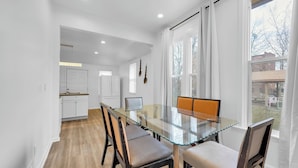 A modern kitchen with white cabinets and a wooden countertop, a dining area with a glass table and chairs, and large windows with curtains. - Spacious 3BR Detroit Apt Sleeps 7 + Wifi (Detroit)