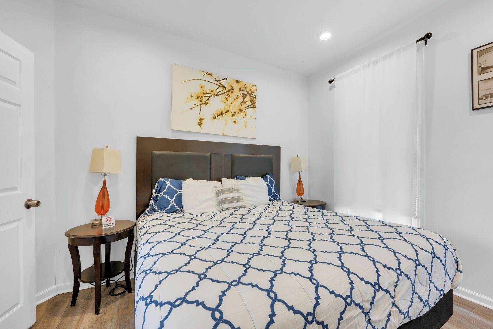 A hotel room with a large bed, two bedside tables, a nightstand, a wall-mounted artwork, and a window with curtains.