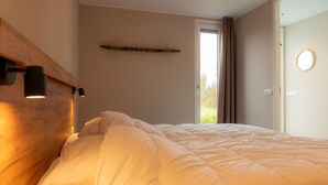 Un dormitorio con cama, cabecera de madera, lámpara de noche, ventana con cortinas y puerta con espejo redondo.