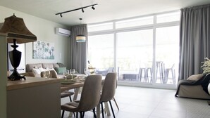 A spacious living area with a dining table, chairs, and a sofa. There is a large window providing natural light and a view outside.