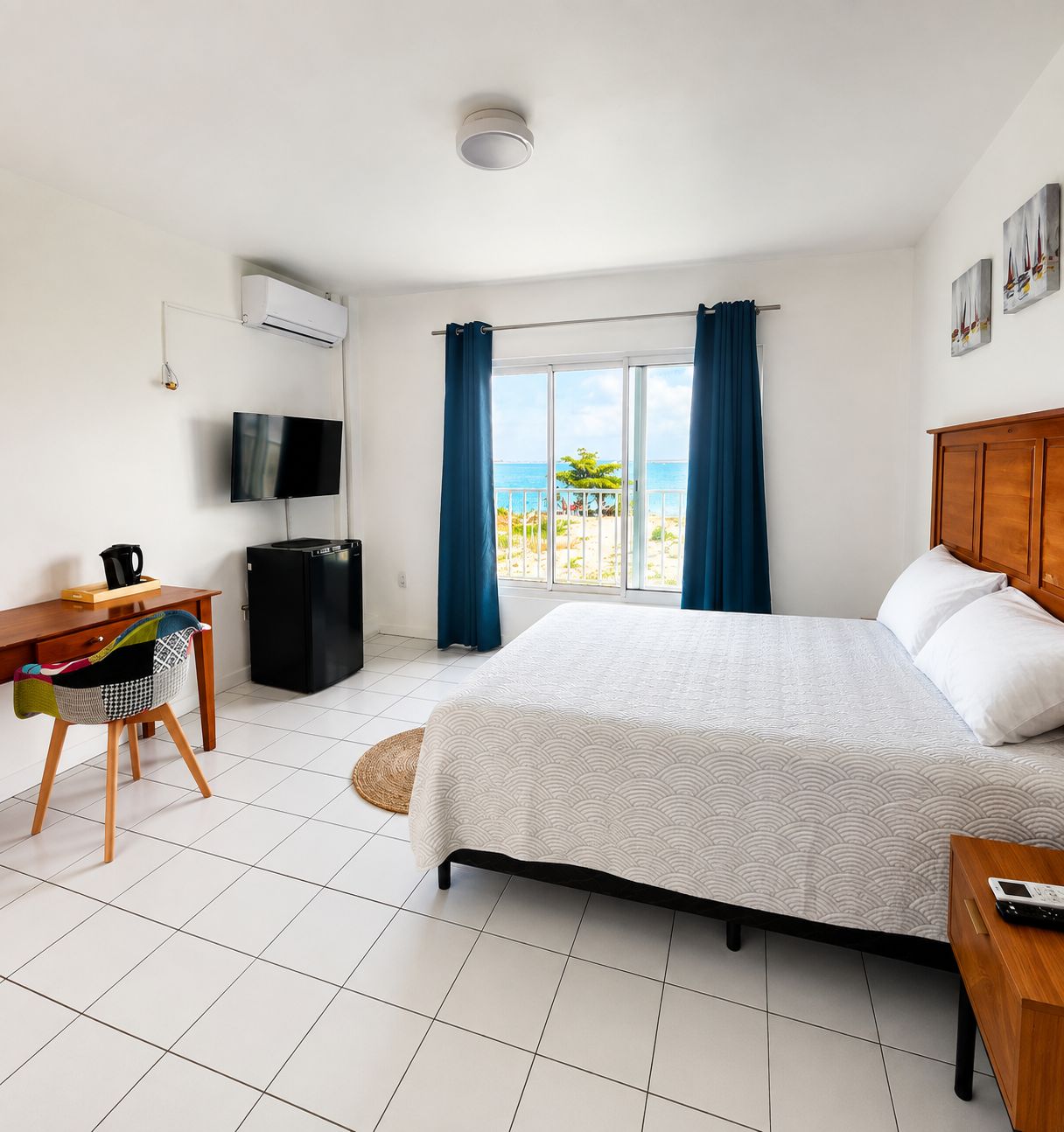 A hotel room with a bed, a desk with a chair, a television, and a view of the beach.