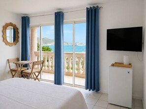 Habitación de hotel con cama, escritorio con sillas, televisor y balcón con cortinas.