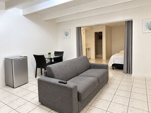 A modern living room with a grey sofa, a small dining table, and a refrigerator. - MIDTOWN MOTEL SXM (Philipsburg)