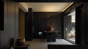 A modern interior with a dark wood panel wall, a dining area with a table and chairs, and a large window providing natural light.