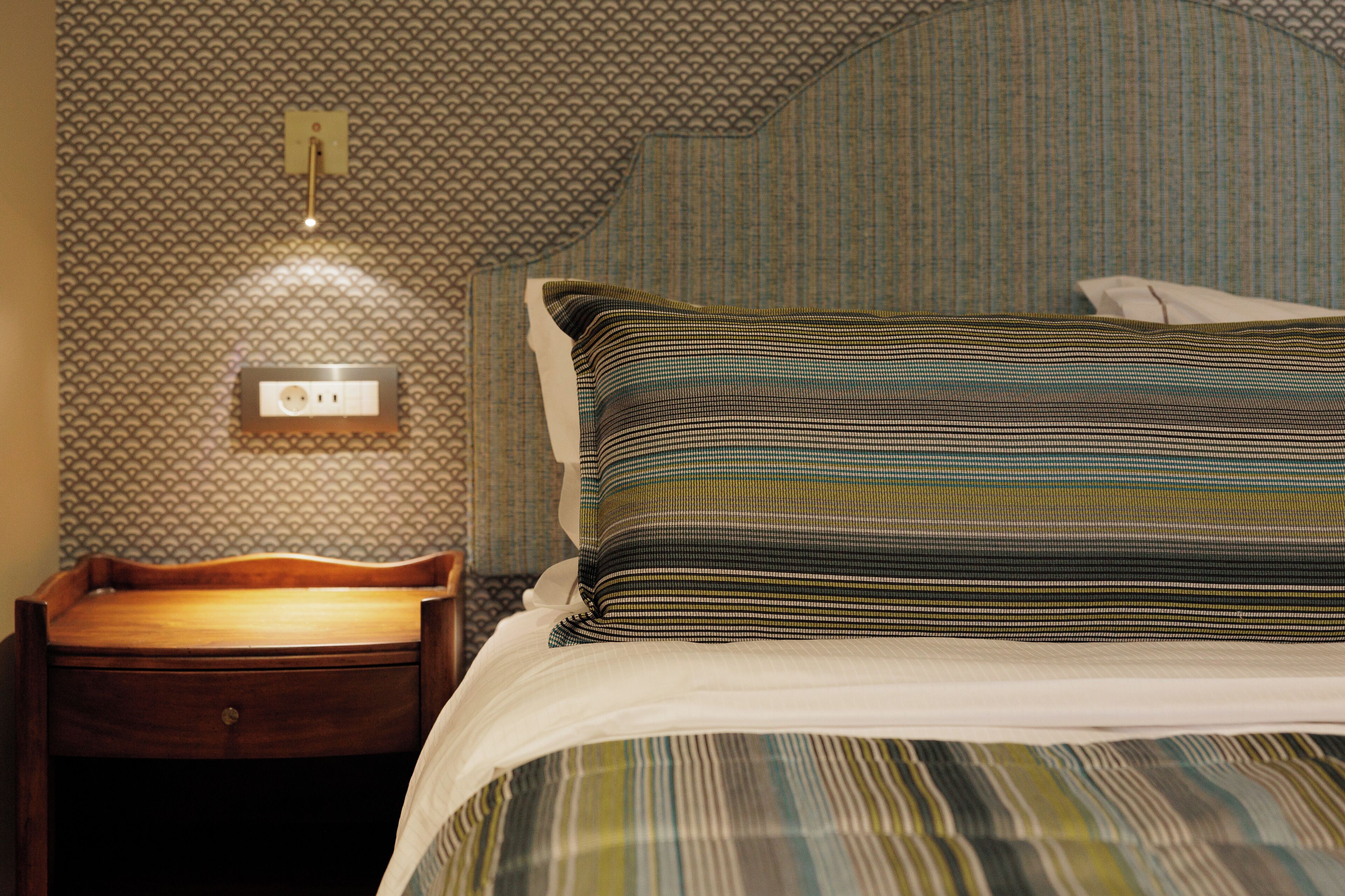 Een net opgemaakt bed met streepjesbeddengoed, een houten nachtkastje met lade en een stopcontact.