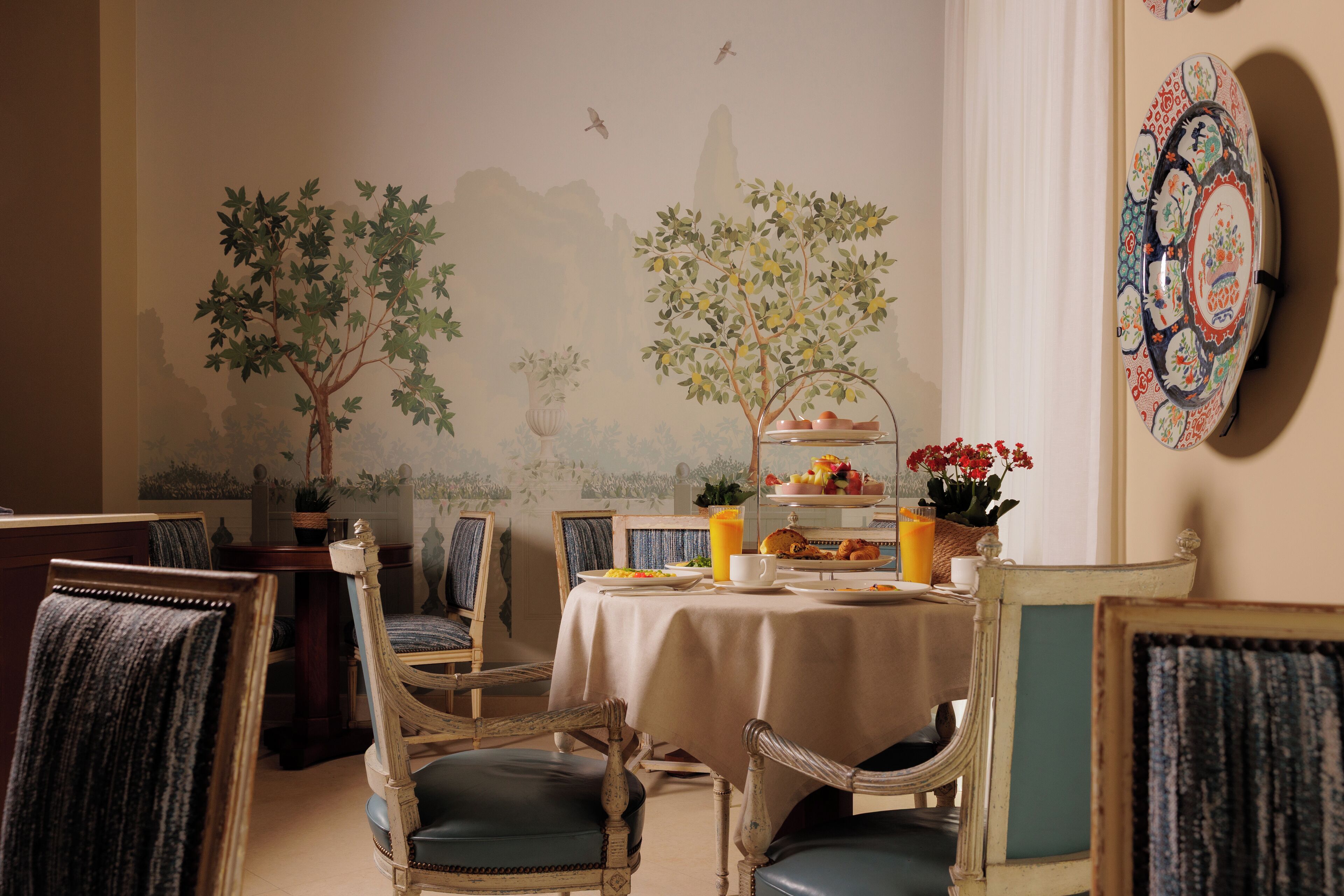 A dining area with a table set for breakfast, chairs, and a mural of trees and birds on the wall.