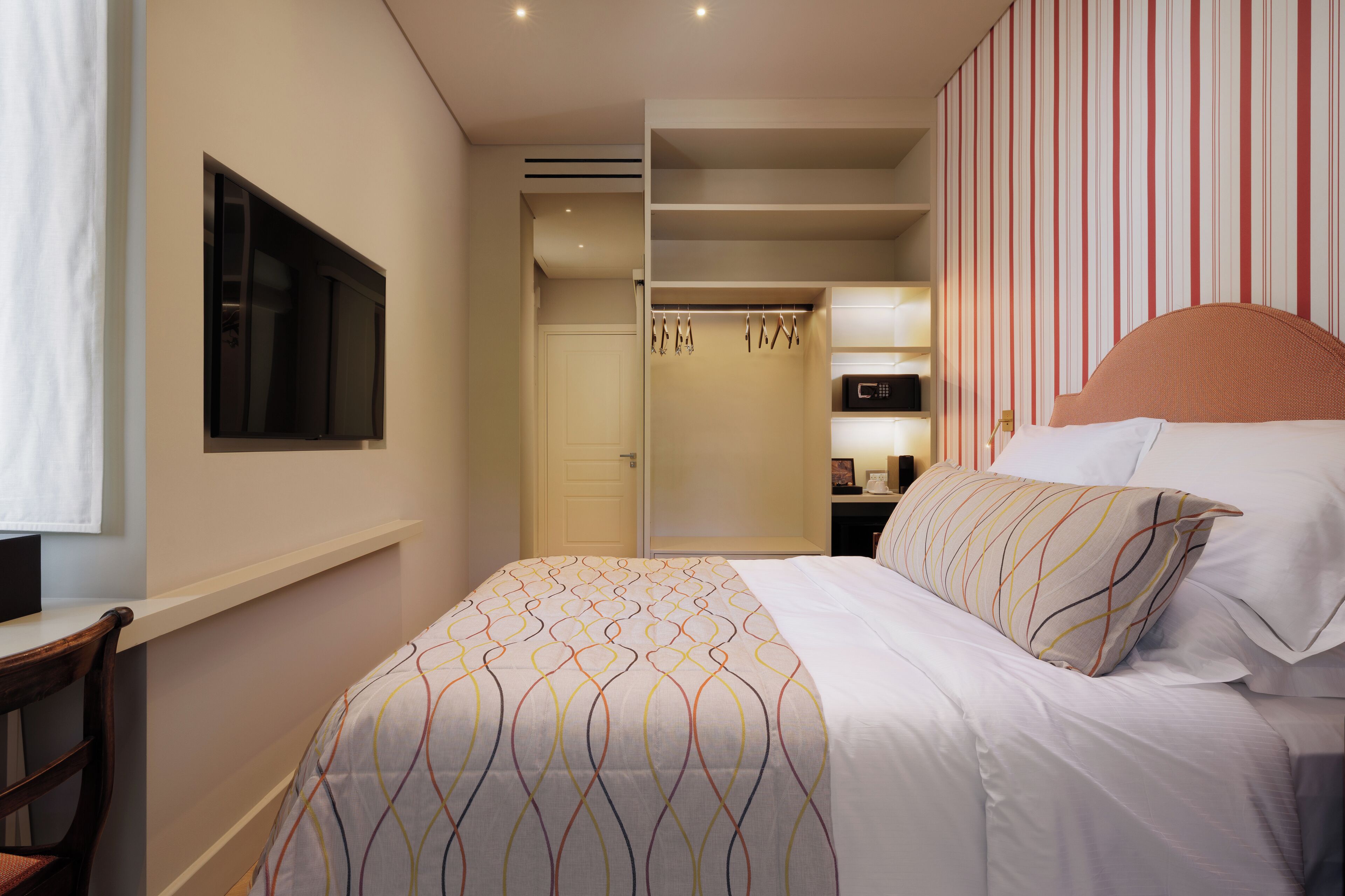 A hotel room with a bed, a TV mounted on the wall, a closet with hanging clothes, and a striped wall.