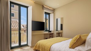 Habitación de hotel con cama, escritorio, silla, televisión y balcón con barandilla.