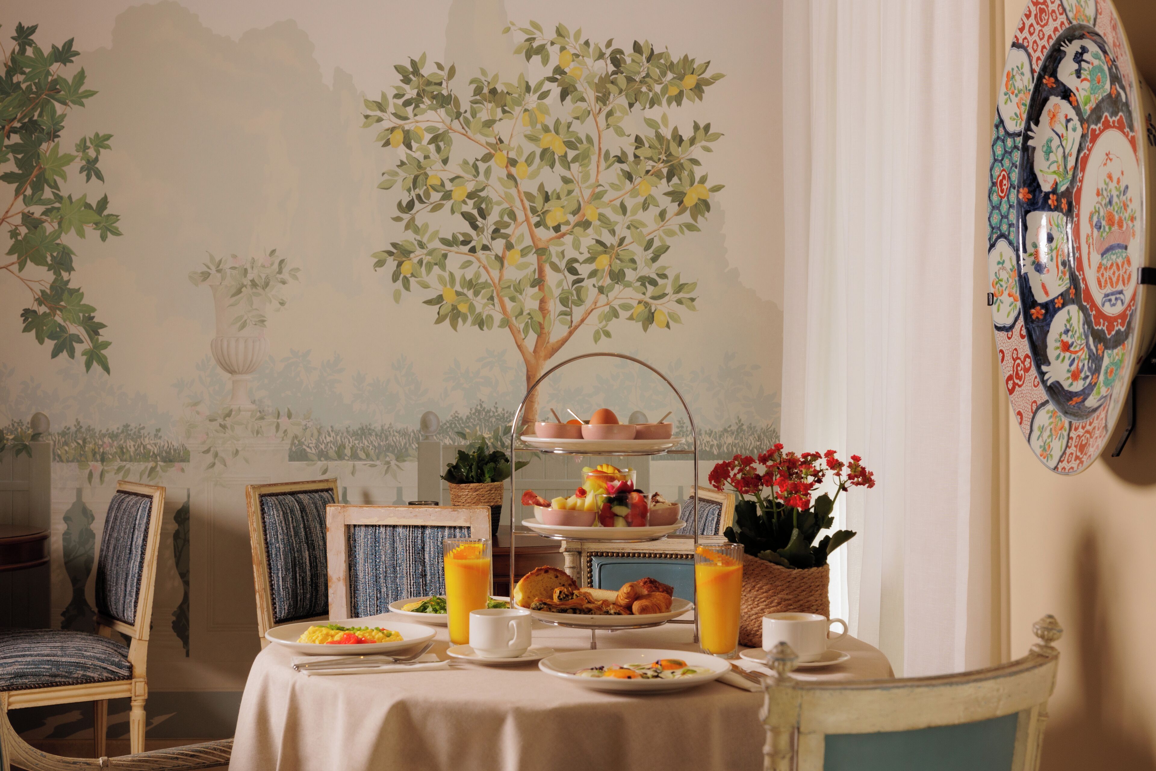 Impostazione colazione con un vassoio a più livelli per frutta e uova, un vaso di fiori e un tavolo con piatti, tazze e una brocca di succo d'arancia.