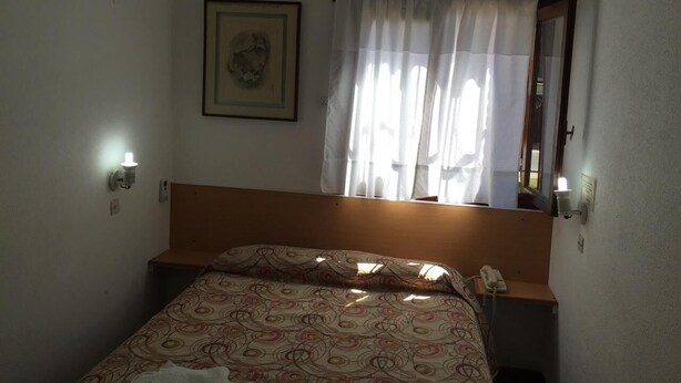 A small, simple room with a bed, a bedside lamp, a wall-mounted air conditioner, a framed picture, and a window with curtains. - Casablanca (Villa Gesell)