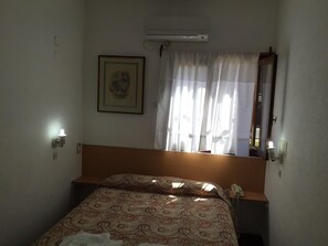 A small, simple room with a bed, a bedside lamp, a wall-mounted air conditioner, a framed picture, and a window with curtains. - Casablanca (Villa Gesell)