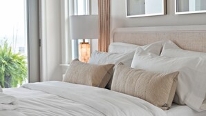 Een netjes opgemaakt bed met witte lakens en meerdere kussens, een bedlamp en een kunstwerk aan de muur.