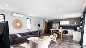 A modern living room with a sofa, armchair, and TV stand. There is a dining area with a table and chairs. The kitchen is visible in the background with cabinets and appliances. - Villa La Rosière, luxury 4-suite villa, private pool & jacuzzi (STE ANNE)