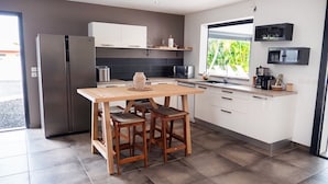 A modern kitchen with a stainless steel refrigerator, wooden table, and bar stools. It features white cabinets, a black backsplash, and a window with a view of greenery. - Villa La Rosière, luxury 4-suite villa, private pool & jacuzzi (STE ANNE)
