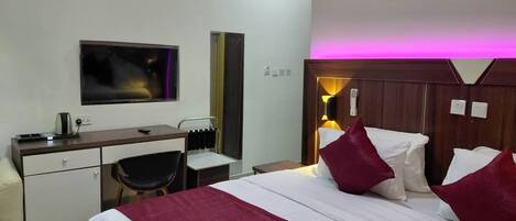 Quarto de hotel com cama, escrivaninha, cadeira e televisão fixada na parede.