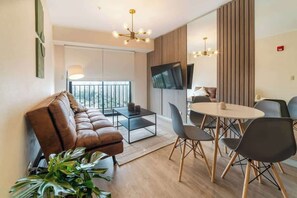 A modern living room with a leather sofa, a dining table with chairs, a TV mounted on the wall, and a large window with a view. - Geko | Modern 1BR Steps to the Sea w/Pool (Barranco)