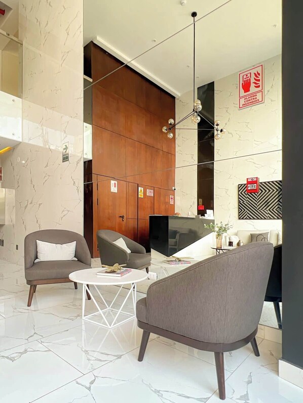 A modern living room with a sofa, armchair, and coffee table, a reception desk, and a fire alarm sign. - Geko | Modern 1BR Steps to the Sea w/Pool (Barranco)