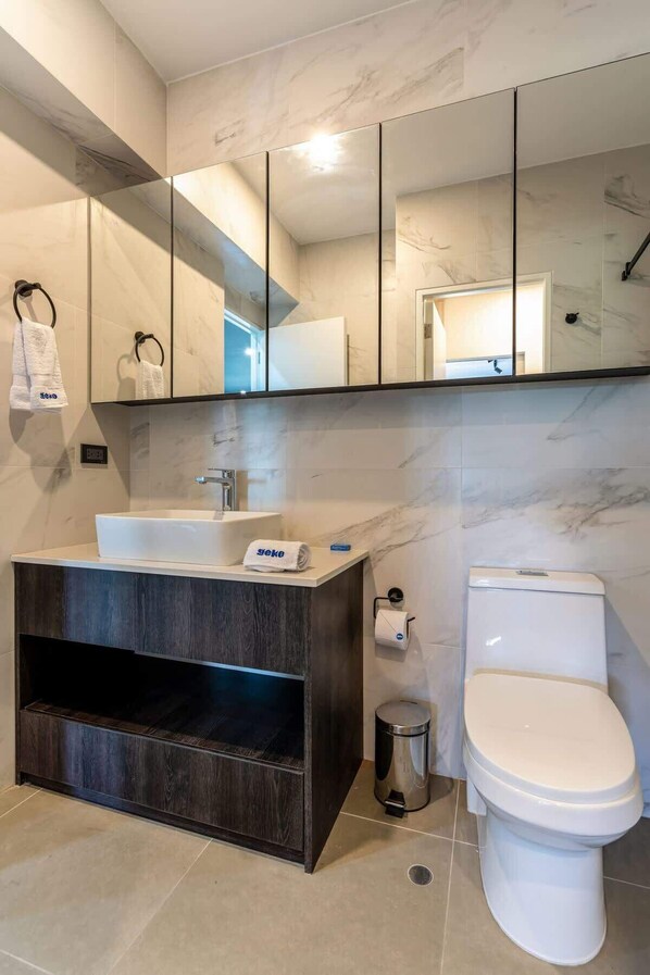 A modern bathroom with a large mirror, a white sink, a dark wood vanity, a toilet, and a trash can.
