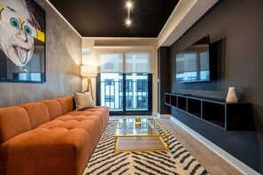 A modern living room with a sofa, a glass coffee table, a patterned rug, a large TV, and a city view through the windows. - Geko | Trendy 1BR w/ Pool in Barranco (Lima)
