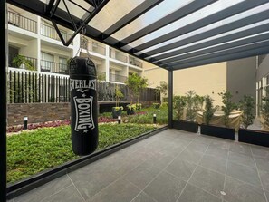 Un espacio al aire libre techado con un saco de boxeo, plantas en macetas y un edificio con balcones al fondo.