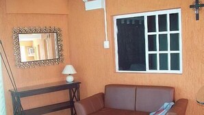 Una habitació amb un sofà de cuir marró, una taula de consola de fusta amb una làmpada, un mirall amb marc decoratiu i una finestra amb persianes blanques.
