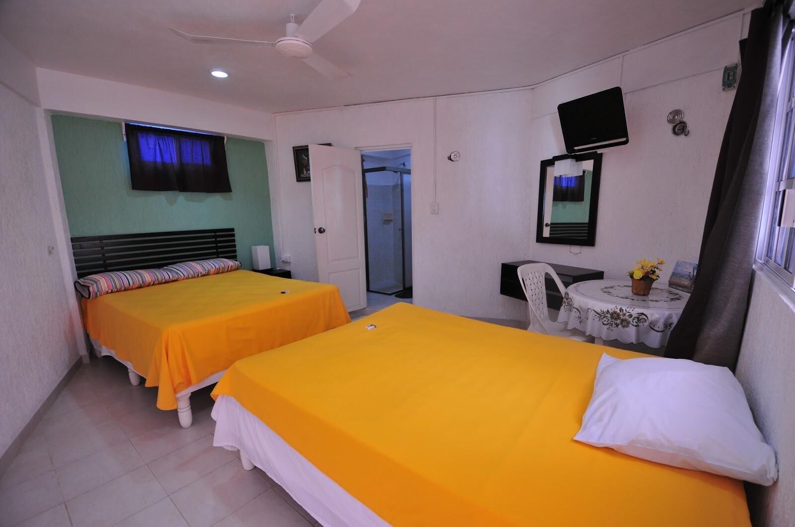 Una habitación de hotel con dos camas, una televisión, una mesita con una silla y una puerta que da a otra habitación.