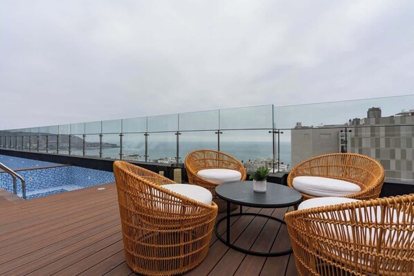 Eine Dachterrasse mit Korbmöbeln, einem kleinen Tisch und Blick auf Stadt und Meer.