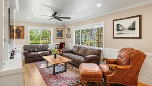 A spacious living room with a brown leather sofa, a wooden coffee table, and a leather armchair. There are large windows with a view of trees and a fence. - 1900's Farmhouse (New Castle)