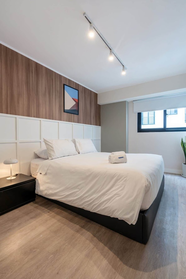 Une chambre moderne avec un grand lit, une table de chevet, une commode, une lampe fixée au mur et une fenêtre donnant sur un beau paysage.