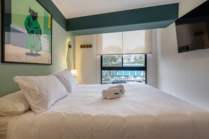 Habitación de hotel con cama, ventana que da a coches aparcados, un cuadro en la pared y un perchero.