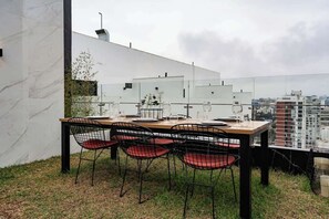 Un espace de restauration en plein air avec une table dressée pour quatre personnes, entourée de chaises dotées de coussins rouges, le tout adossé à un mur blanc offrant une vue sur le paysage urbain.